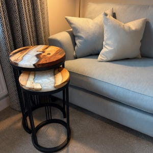 May include: A set of two nesting tables featuring circular tops made from natural wood slabs with a central inlay of translucent white resin. The tables are supported by thin, black metal frames with circular bases, providing a stable structure.