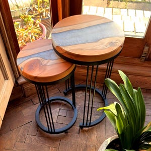 May include: Two wooden side tables with round tops and black metal legs. The table tops are made of light brown wood with a gray resin river design. The tables have a modern, minimalist design.