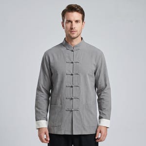 May include: A long-sleeved grey shirt featuring a mandarin collar and a front closure with five fabric knot buttons. The garment includes two lower patch pockets and contrasting white cuffs at the end of each sleeve, providing a clean look.