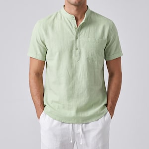 Puede incluir: Una camisa de color verde salvia claro de manga corta con cuello mao, una tapeta de botones parcial con cuatro botones visibles y un bolsillo en el pecho en el lado izquierdo. La camisa tiene un dobladillo recto y mangas cortas con vuelta.