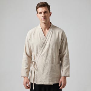 May include: A beige wrap-style jacket with long sleeves, a V-neckline, and a side tie closure. The garment features a single patch pocket on the lower left side and is constructed from a textured fabric with visible weave details throughout the piece.