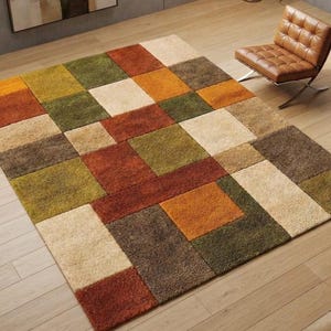 Könnte beinhalten: Ein rechteckiger Teppich mit einem geometrischen Muster aus großen, unterschiedlich großen Quadraten und Rechtecken in den Farben Olivgrün, gebranntes Orange, Creme und Dunkelbraun. Der Teppich hat eine strukturierte Oberfläche und liegt auf einem hellen Holzboden.
