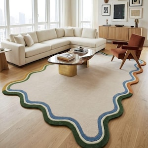 Op de afbeelding: Een groot, cr&egrave;mekleurig vloerkleed met een onregelmatige, golvende, organische vorm. De rand is voorzien van gelaagde, golvende banden in donkergroen, lichtgroen, blauw en oranje, wat een duidelijk contrast vormt met het neutrale midden.