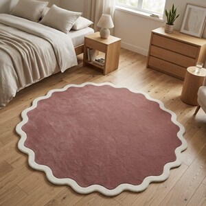 May include: A large, circular area rug featuring a solid dusty rose center and a thick, cream-colored scalloped border. The rug has a soft, textured pile surface and is positioned flat on a light-toned wooden floor in a bedroom setting.