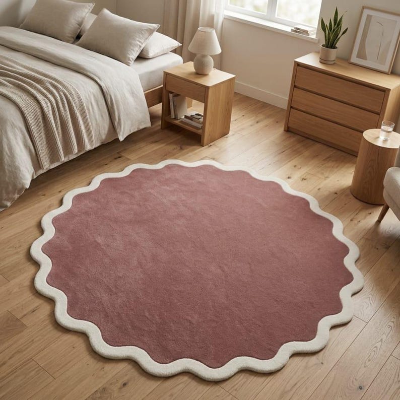 May include: A large, circular area rug featuring a solid dusty rose center and a thick, cream-colored scalloped border. The rug has a soft, textured pile surface and is positioned flat on a light-toned wooden floor in a bedroom setting.