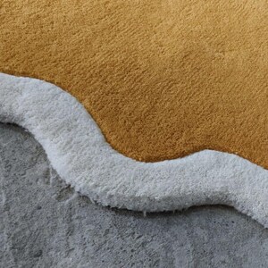 May include: A tufted rug featuring a large, solid mustard yellow section bordered by a thick, wavy white trim. The rug has a dense, plush pile texture and an irregular, organic shape that follows the curves of the white border edge.