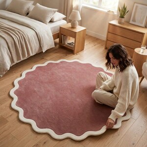 May include: A large, circular area rug featuring a solid dusty rose center and a thick, cream-colored scalloped border. The rug has a soft, plush texture and is laid flat on a light-toned wooden floor, showcasing its distinct wavy edge design.