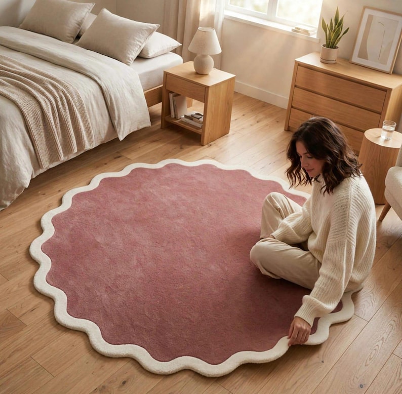 May include: A large, circular area rug featuring a solid dusty rose center and a thick, cream-colored scalloped border. The rug has a soft, plush texture and is laid flat on a light-toned wooden floor, showcasing its distinct wavy edge design.