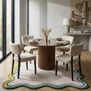 Op de afbeelding: Een ronde eettafel met een geribbelde voet van donker hout en een glazen blad, omringd door vier stoelen bekleed met cr&egrave;mekleurige, gestructureerde stof met donkerhouten poten, geplaatst op een groot vloerkleed met een golvende, meerkleurige rand.