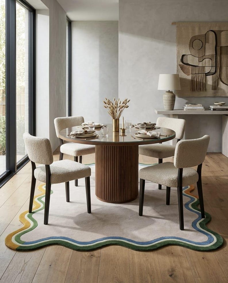 Op de afbeelding: Een ronde eettafel met een geribbelde voet van donker hout en een glazen blad, omringd door vier stoelen bekleed met cr&egrave;mekleurige, gestructureerde stof met donkerhouten poten, geplaatst op een groot vloerkleed met een golvende, meerkleurige rand.