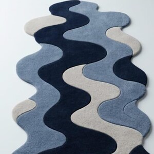 Pode incluir: Um tapete com formato org&acirc;nico ondulado e um padr&atilde;o em camadas de curvas entrela&ccedil;adas. O design consiste em blocos de cores s&oacute;lidas em azul-marinho profundo, azul-ard&oacute;sia suave e bege claro, criando um fluxo visual cont&iacute;nuo e fluido.