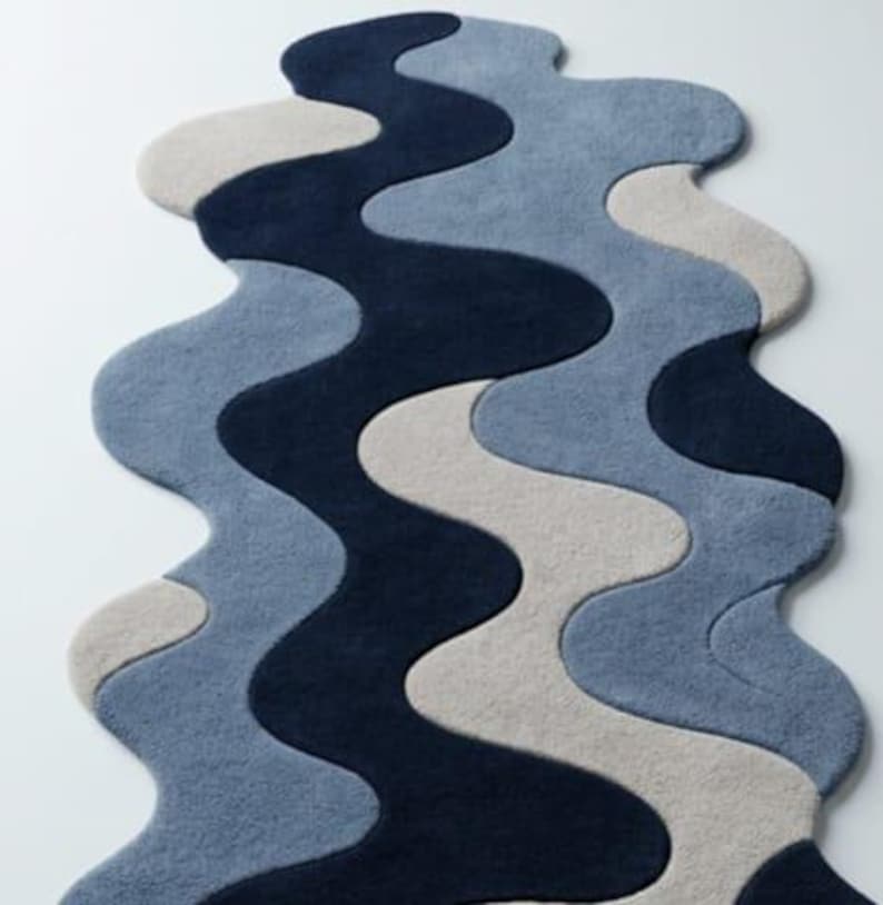 Pode incluir: Um tapete com formato org&acirc;nico ondulado e um padr&atilde;o em camadas de curvas entrela&ccedil;adas. O design consiste em blocos de cores s&oacute;lidas em azul-marinho profundo, azul-ard&oacute;sia suave e bege claro, criando um fluxo visual cont&iacute;nuo e fluido.