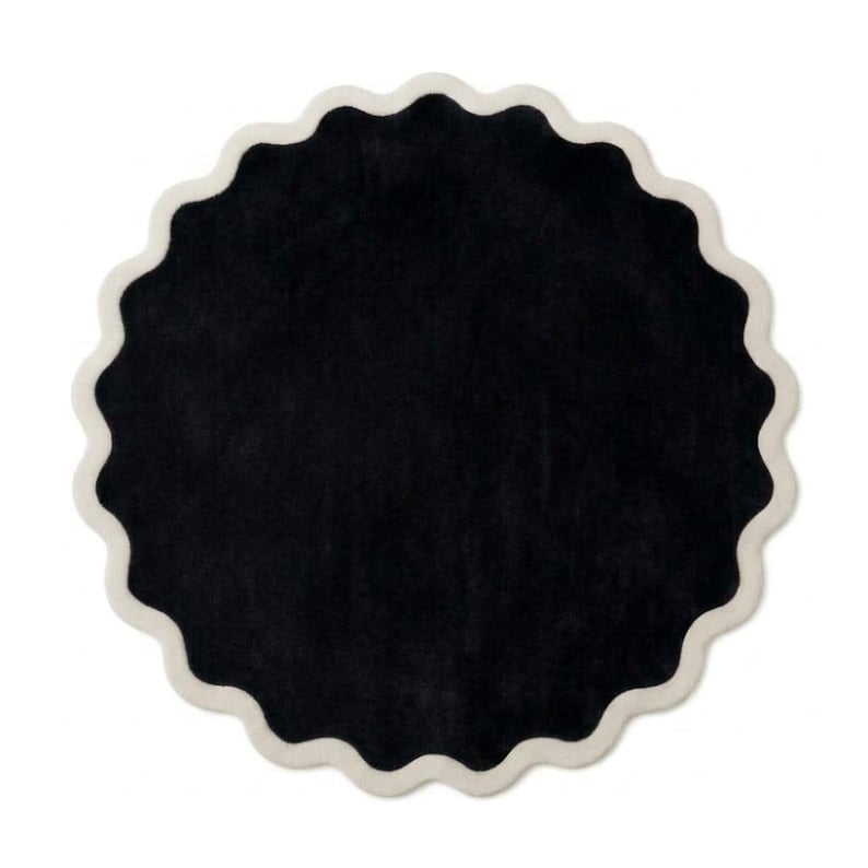 May include: A circular rug featuring a solid black center and a scalloped edge finished with a thick, cream-colored border. The design is symmetrical, with a soft, textured surface that creates a distinct contrast between the dark interior and light trim.