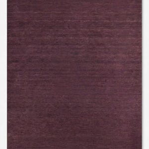 May include: A rectangular area rug in a deep, solid plum purple color. The surface features a consistent, fine-grained horizontal texture throughout, creating a subtle variation in light and shadow across the flat, woven fabric construction.
