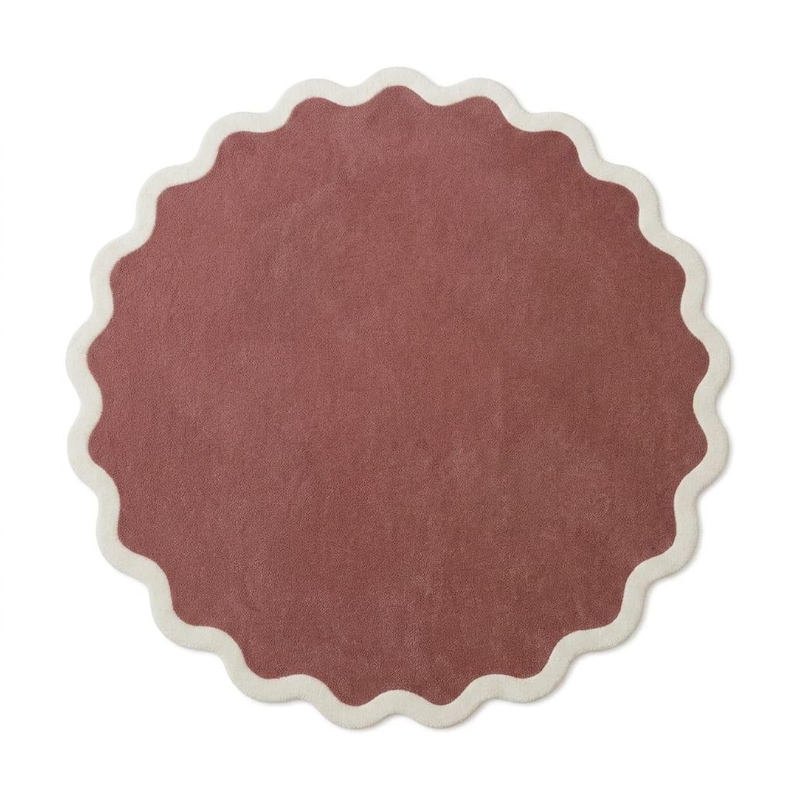 May include: A circular rug featuring a solid dusty rose center and a scalloped edge finished with a thick cream-colored border. The textile surface has a uniform, soft texture throughout, creating a distinct contrast between the muted pink and the light neutral trim.