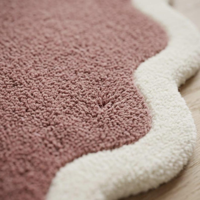 May include: A tufted rug featuring a dusty rose center with a thick, scalloped cream-colored border. The rug has a dense, textured pile surface with a soft, looped appearance, creating a distinct contrast between the two colors and shapes.