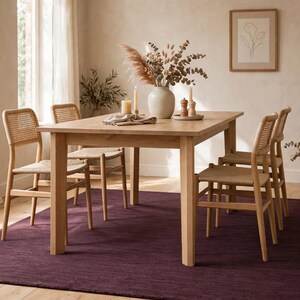 May include: A light wood rectangular dining table with four matching chairs featuring woven cane backrests and seats. The table is set with a ceramic vase containing dried foliage, two candles, and a small wooden salt and pepper shaker set.