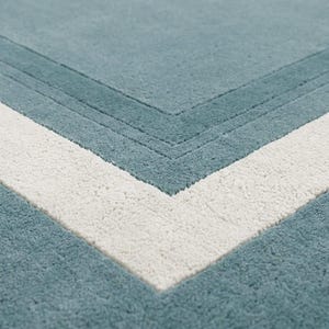 Könnte beinhalten: Ein flauschiger, getufteter Teppich mit einer einfarbigen, staubblauen Oberfläche und einer markanten weißen V-förmigen Bordüre. Das Design weist vertiefte, eingearbeitete parallele Linien auf, die dem Winkel folgen und Struktur erzeugen.