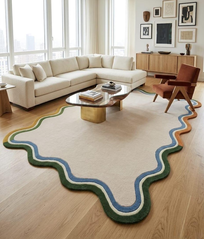Op de afbeelding: Een groot, cr&egrave;mekleurig vloerkleed met een onregelmatige, golvende, organische vorm. De rand is voorzien van gelaagde, golvende banden in donkergroen, lichtgroen, blauw en oranje, wat een duidelijk contrast vormt met het neutrale midden.