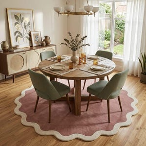 May include: A round wooden dining table with four sage green upholstered chairs, set on a large circular dusty rose rug with a cream scalloped border. The table is arranged with place settings, amber glassware, and a central vase with greenery.