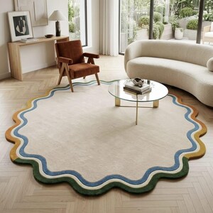 Op de afbeelding: Een groot, rond vloerkleed met een golvende, geschulpte rand. Het midden is effen lichtbeige, omringd door concentrische, golvende banden in blauw, cr&egrave;me en een mix van mosterdgeel en donkergroene segmenten langs de buitenrand.