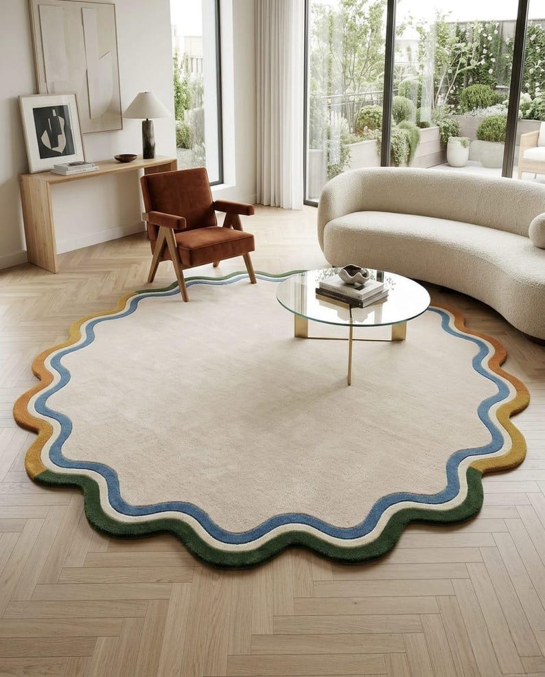Op de afbeelding: Een groot, rond vloerkleed met een golvende, geschulpte rand. Het midden is effen lichtbeige, omringd door concentrische, golvende banden in blauw, cr&egrave;me en een mix van mosterdgeel en donkergroene segmenten langs de buitenrand.