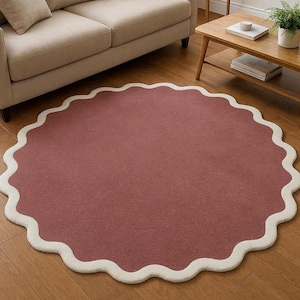 May include: A circular rug featuring a solid dusty rose center and a thick, scalloped cream-colored border. The rug has a soft, textured pile surface and is laid flat on a wooden floor, showcasing a distinct wavy edge design throughout its perimeter.