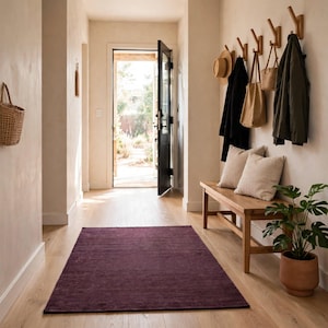 May include: A rectangular deep purple area rug centered on a light wood floor in an entryway. The space features a wooden bench with two beige pillows, a row of wall-mounted coat hooks holding bags and jackets, and a potted green plant.