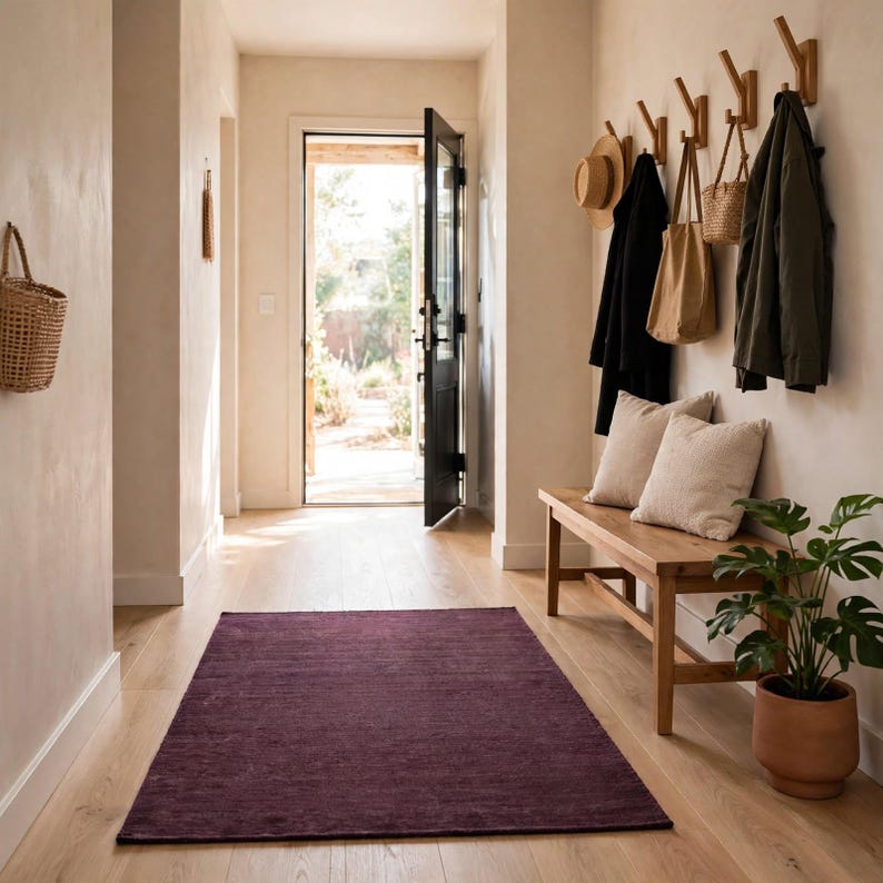 May include: A rectangular deep purple area rug centered on a light wood floor in an entryway. The space features a wooden bench with two beige pillows, a row of wall-mounted coat hooks holding bags and jackets, and a potted green plant.