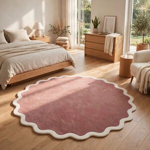 May include: A circular area rug featuring a solid dusty rose center and a thick, scalloped cream-colored border. The rug is placed on a light-toned wooden floor in a bedroom setting, showcasing a distinct wavy edge design throughout its perimeter.