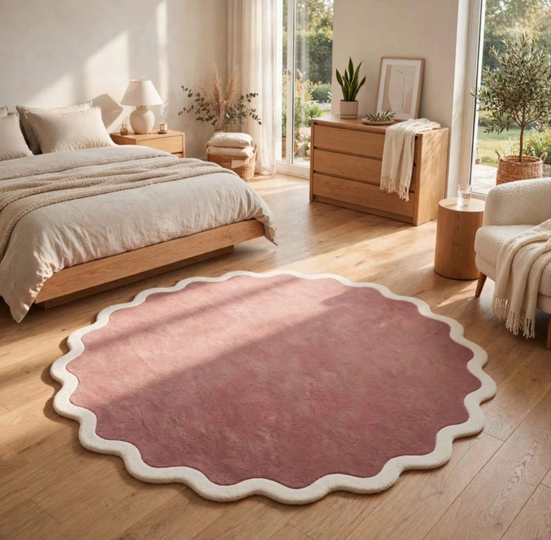 May include: A circular area rug featuring a solid dusty rose center and a thick, scalloped cream-colored border. The rug is placed on a light-toned wooden floor in a bedroom setting, showcasing a distinct wavy edge design throughout its perimeter.