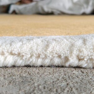 May include: A close-up view of a rug edge showing a thick, white, tufted pile border that transitions into a smooth, solid mustard yellow surface. The fibers are densely packed, revealing the structural construction of the textile material.