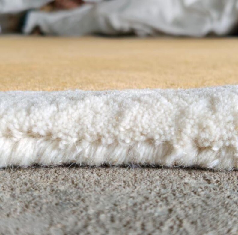 May include: A close-up view of a rug edge showing a thick, white, tufted pile border that transitions into a smooth, solid mustard yellow surface. The fibers are densely packed, revealing the structural construction of the textile material.