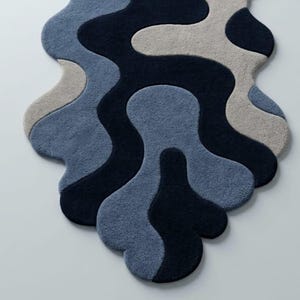 Pode incluir: Um tapete com formato org&acirc;nico irregular e textura tufada. O design consiste em padr&otilde;es fluidos e interligados em tons de azul-marinho escuro, azul m&eacute;dio e bege claro, criando uma apar&ecirc;ncia abstrata e em camadas.