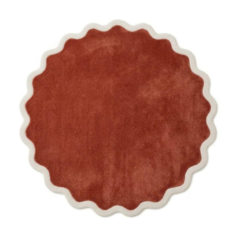 May include: A circular rug featuring a solid terracotta-colored center with a soft, textured surface. The rug is framed by a thick, scalloped border in a contrasting cream color, creating a distinct wavy edge around the entire perimeter.