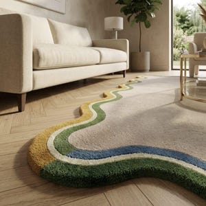 Op de afbeelding: Een vloerkleed met een onregelmatige, golvende vorm met een groot beige middenstuk, omrand door concentrische, golvende strepen in mosterdgeel, cr&egrave;me, saliegroen en diepblauw. Het kleed heeft een dikke, getufte textuur en ligt op een houten vloer.