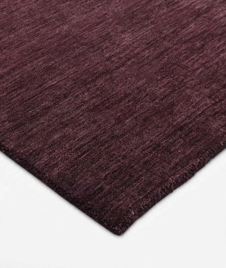 May include: A deep burgundy area rug featuring a dense, textured pile with a subtle horizontal grain pattern. The surface displays variations in shade and light reflection across the fibers, creating a rich, monochromatic appearance throughout the rug.
