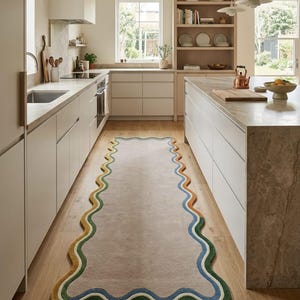 Op de afbeelding: Een lange, rechthoekige loper met een golvende, geschulpte rand. Het midden is effen lichtbeige, omzoomd door concentrische, golvende strepen in tinten donkergroen, lichtgroen, blauw en oranje, geplaatst op een lichte houten keukenvloer.