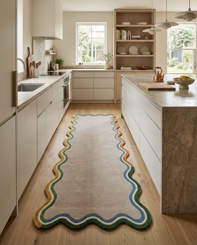 Op de afbeelding: Een lange, rechthoekige loper met een golvende, geschulpte rand. Het midden is effen lichtbeige, omzoomd door concentrische, golvende strepen in tinten donkergroen, lichtgroen, blauw en oranje, geplaatst op een lichte houten keukenvloer.