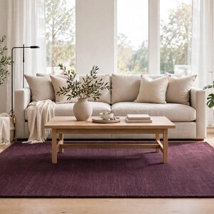 May include: A rectangular area rug in a deep, solid plum purple color with a subtle, low-pile texture. The rug is positioned on a light-toned wooden floor, providing a bold, dark contrast to the neutral-colored furniture and decor items placed above it.