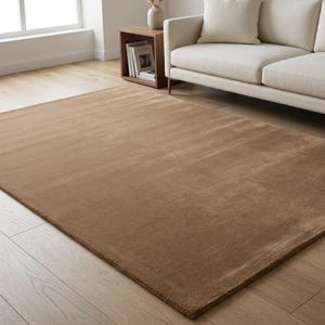 May include: A large, rectangular brown rug with a soft, plush texture. The rug is placed on a light-colored hardwood floor, with a white sofa and wooden side table in the background. The rug adds warmth and comfort to the living space.