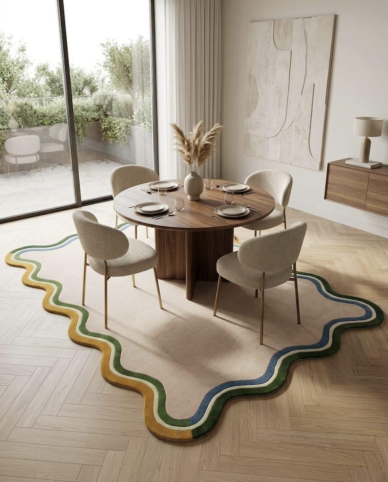 Op de afbeelding: Een groot, onregelmatig gevormd vloerkleed met een golvende, geschulpte rand met concentrische banden in mosterdgeel, bosgroen en marineblauw. Het kleed is beige en ligt onder een ronde houten eettafel met vier cr&egrave;mekleurige stoelen.