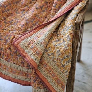 May include: A floral patterned quilt in shades of orange, yellow, and gray, draped over a wooden chair. The quilt features a detailed floral design and a contrasting border in a darker shade of orange. The overall aesthetic is warm and inviting.