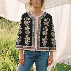 Può includere: Giacca nera ricamata con motivi floreali color crema e beige. La giacca ha un bordo con frange bianche e viene indossata con jeans blu. Lo sfondo presenta erba verde alta e un lenzuolo bianco.