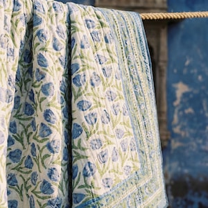 Op de afbeelding: Een witte quilt met een blauw bloemenpatroon en groene stelen hangt aan een touw. De quilt heeft een blauwe rand met een herhalend bloemenontwerp. De achtergrond is een blauwe muur met textuur.