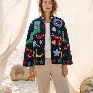Pode incluir: Casaco azul marinho com desenhos bordados coloridos, incluindo botas, cobras, estrelas e um sol. O casaco tem gola e punhos brancos. A modelo usa um top branco e calças bege.