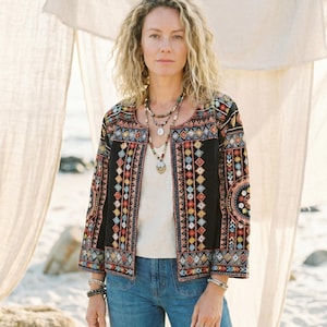 May include: A black jacket with colourful embroidered geometric patterns. The jacket has long sleeves and is worn over a white top and blue jeans. The model is wearing multiple necklaces and bracelets.