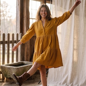 May include: Mustard yellow tiered dress with long sleeves and a V-neck. The dress is paired with brown cowboy boots. The background features a rustic wooden structure and a neutral-colored backdrop.
