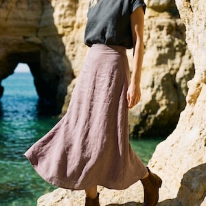 Op de afbeelding: Een persoon draagt een donkergrijze top met korte mouwen en een lange, vloeiende mauve rok. Ze draagt bruine enkellaarsjes. De achtergrond toont een rotsachtige klif en turquoise water.