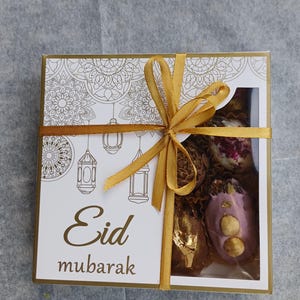 Puede incluir: Una caja de regalo blanca con detalles dorados y una cinta dorada, con la inscripción "Eid Mubarak". Contiene dulces, algunos cubiertos de chocolate y con nueces. El frente tiene un diseño decorativo con faroles.